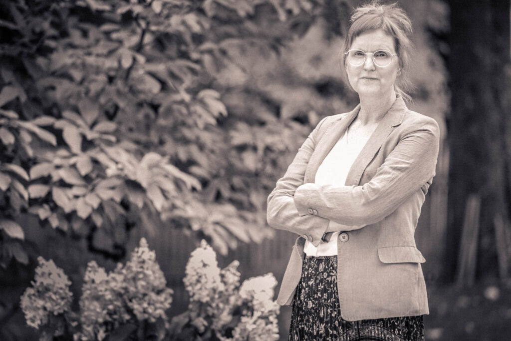 Christine Bethke Trauerrednerin München stehend neben Forsythie mit geschränkten Armen schwarz weiß - oficiante funeraria Múnich - officiante funéraire Munich
