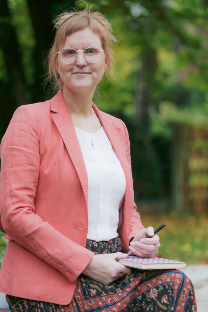 Christine Bethke Trauerrednerin für individuelle Trauerfeiern München sitzend im Garten auf schwarzer Steinbank mit Block und Stift in der Hand und roten Rosen im Hintergrund oficiante funeraria Múnich officiante funéraire Munich